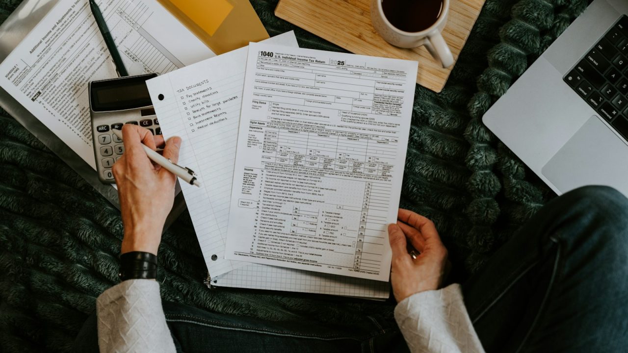 A person reviewing paper financial documents at a desk with a calculator and a laptop